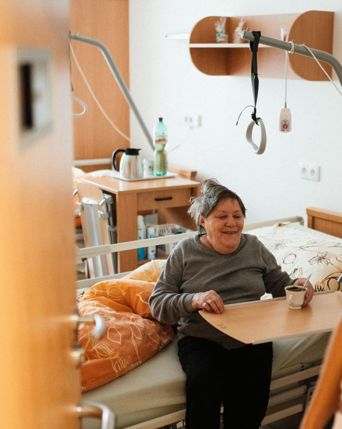 Senhora idosa sentada na beira do leito hospitalar, sorrindo, com uma xícara de café na mão