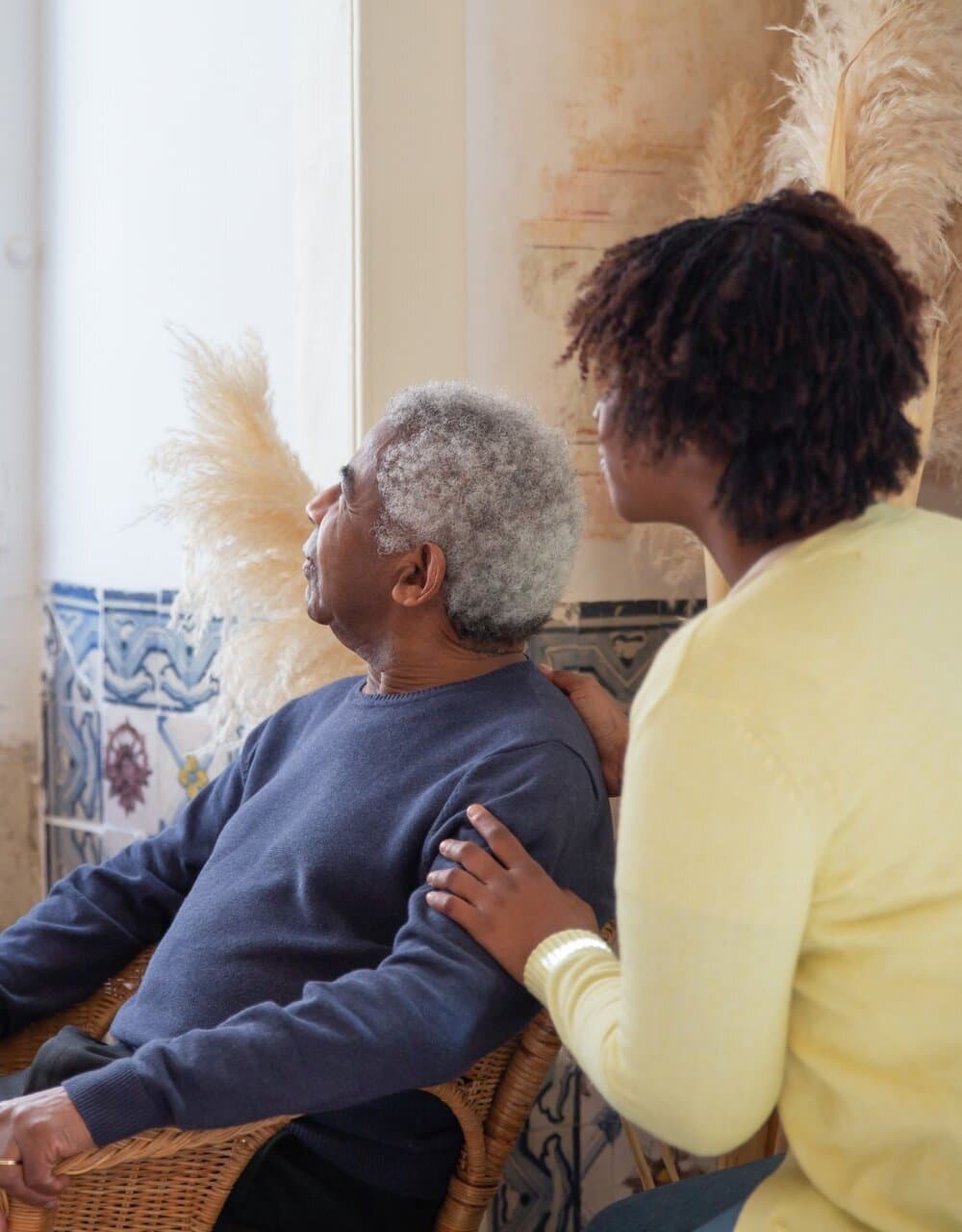 Senhor idoso sentado em uma poltrona de vime em casa, com uma cuidadora ao lado apoiando a mão em seu ombro